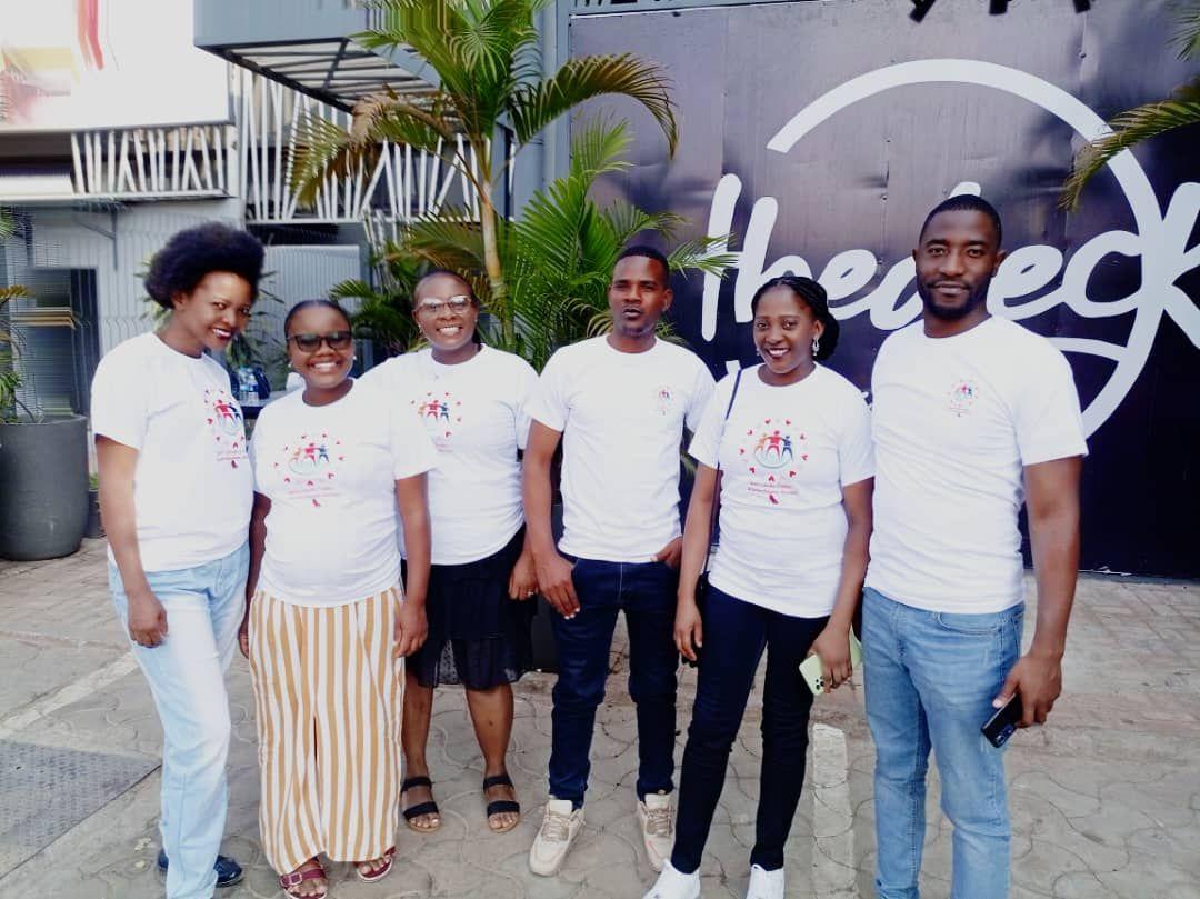 Group of six people in matching white t-shirts smiling outdoors near a building and plants.