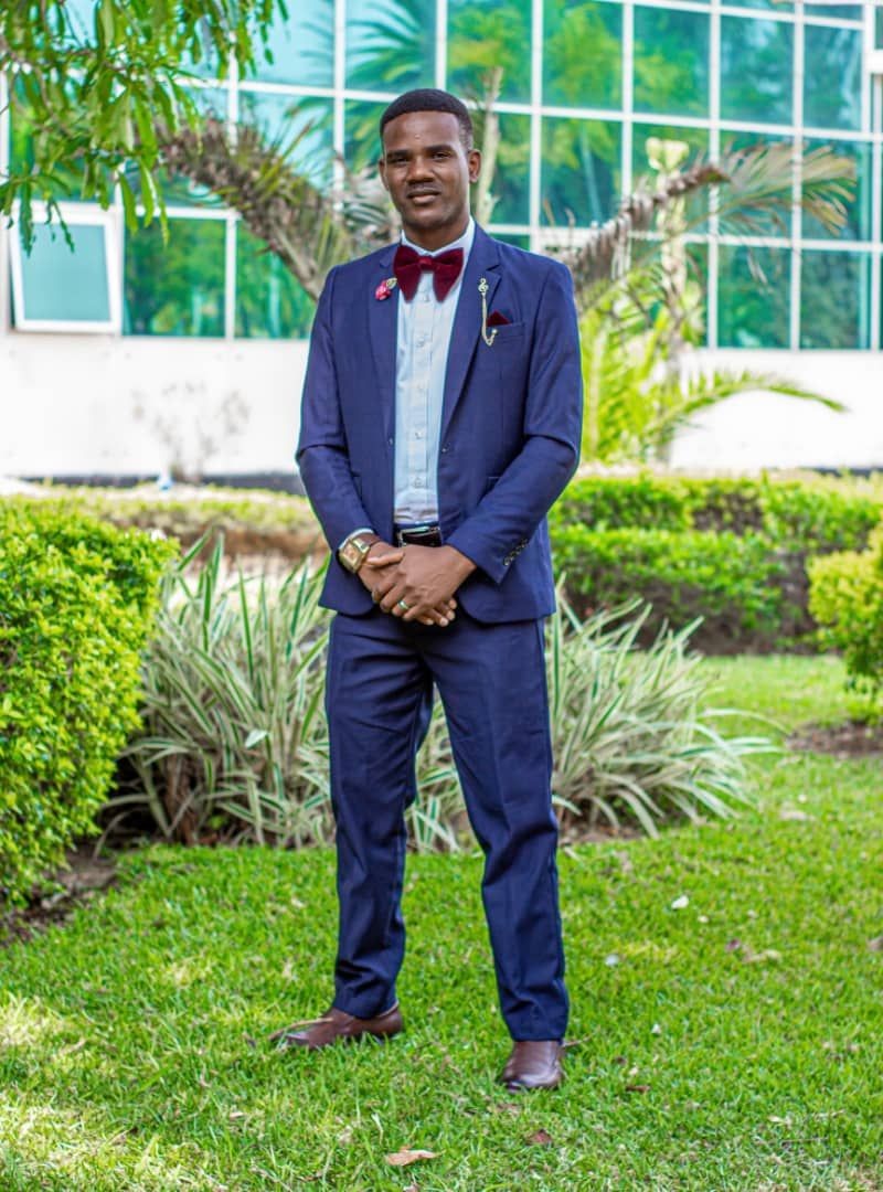 Man in blue suit with red bow tie standing on grass in front of glass building.