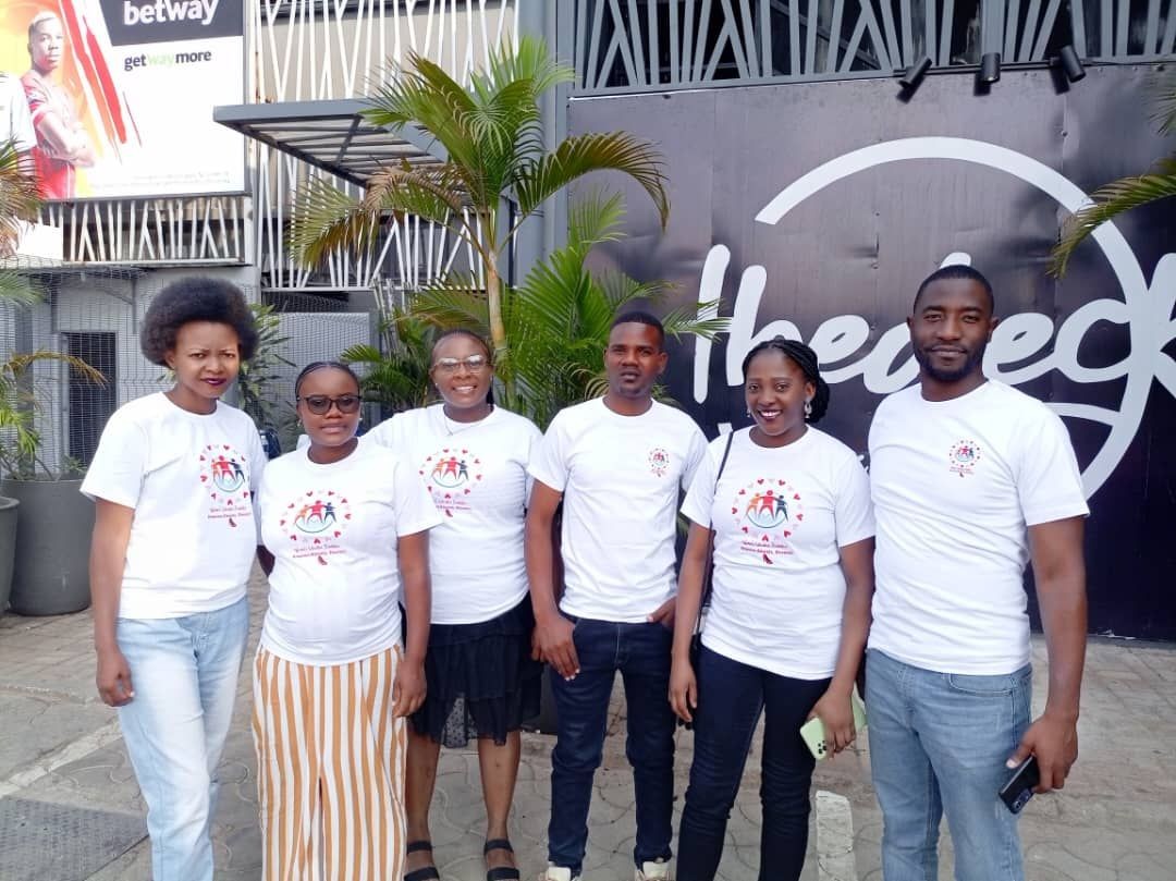 Group of six people in matching white T-shirts posing outdoors near a sign and plants.