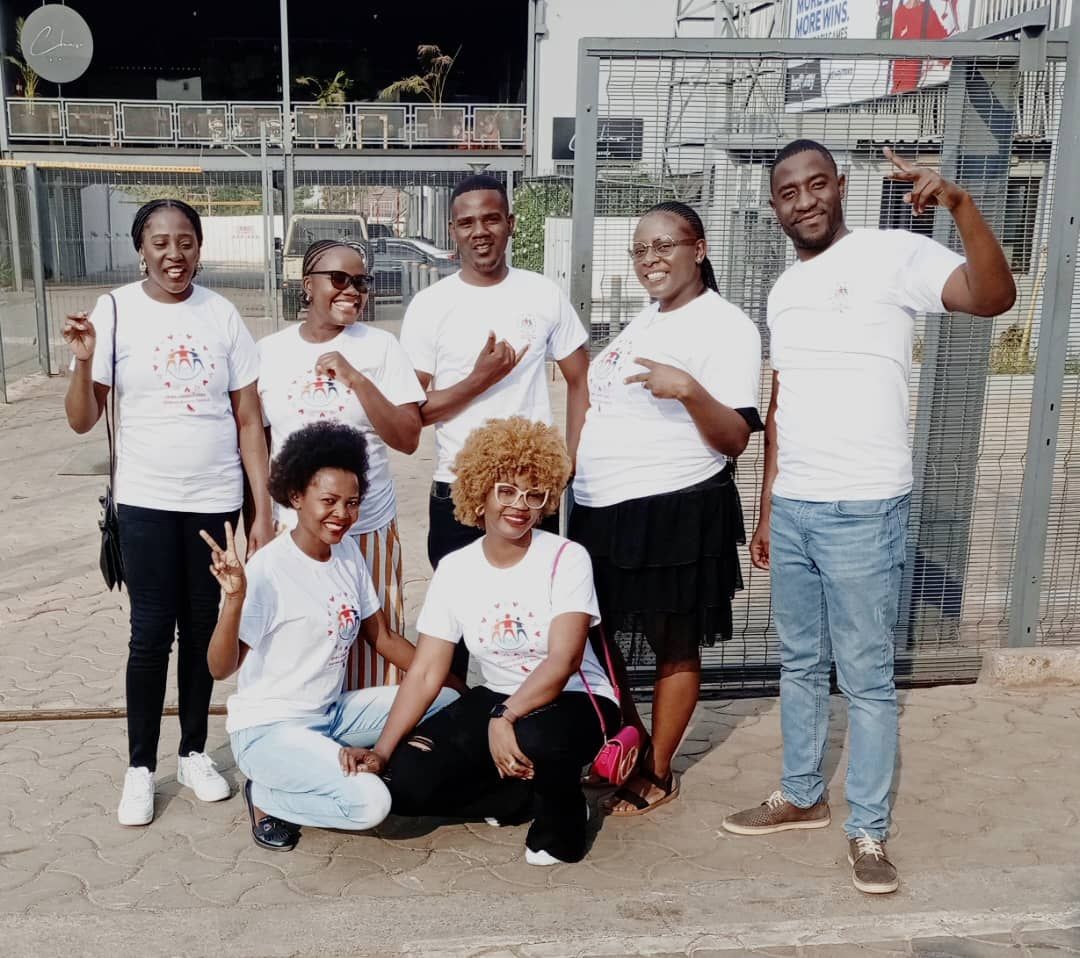 Group of seven people in white shirts posing outdoors, some kneeling and smiling.