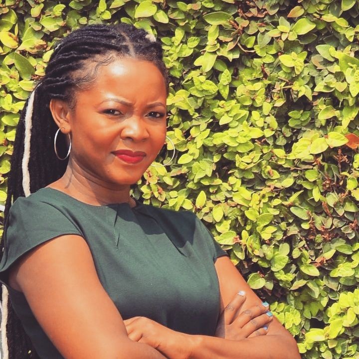 Person with braided hair stands confidently in front of a lush green hedge.