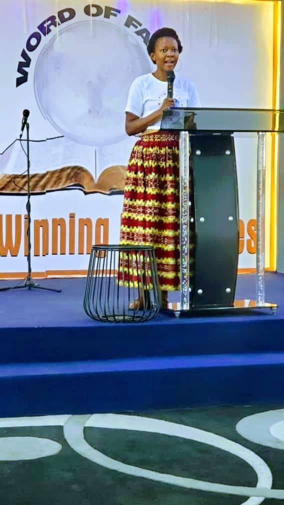 Woman speaking at a podium on a stage, with a microphone and large open book backdrop.