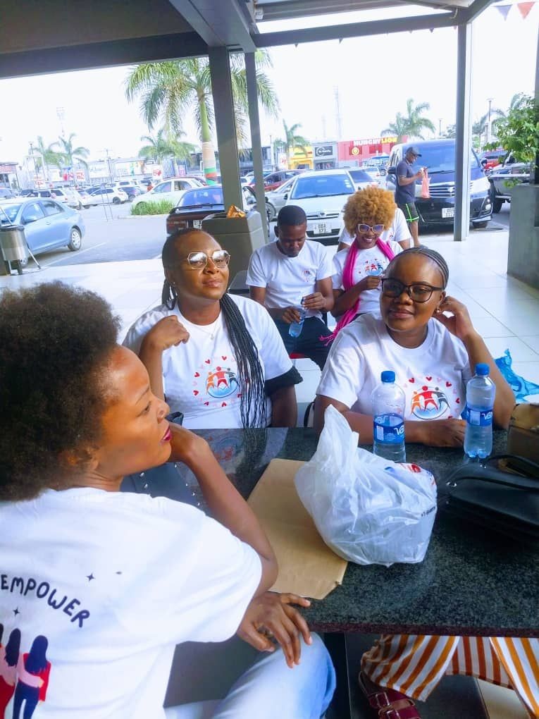 Group of people wearing white T-shirts sitting at an outdoor table near parked cars and palm trees.